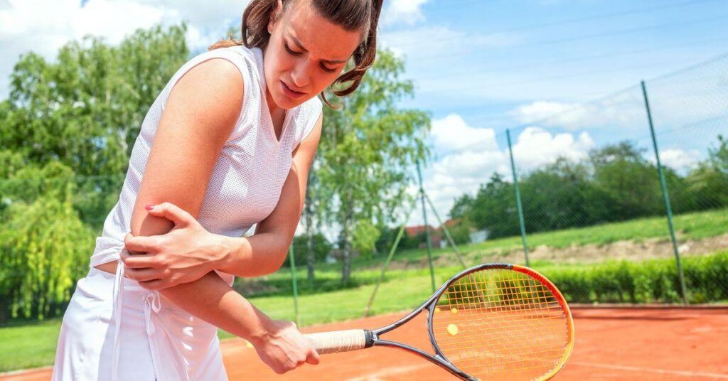 A female tennis player in a white outfit clutches her elbow in discomfort while standing on a clay court, her racket still in hand. Strengthening and stretching with the Best Tennis Elbow Exercises can help prevent and recover from such injuries.