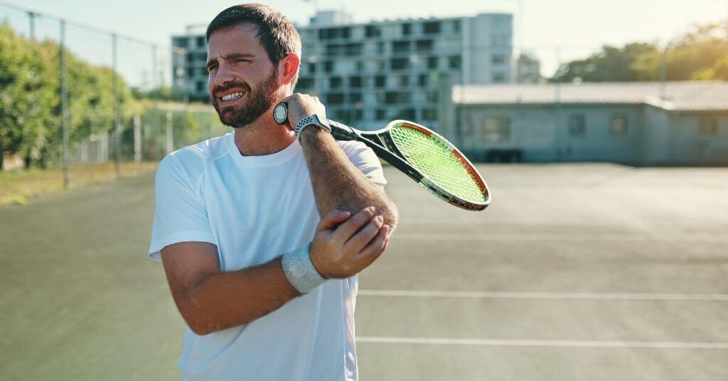 A male tennis player in a white shirt grimaces in pain while holding his elbow on an outdoor court. Overuse injuries like tennis elbow can be treated with rest, physical therapy, and the Best Tennis Elbow Exercises.