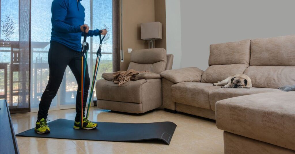 A person in a blue tracksuit performs resistance band exercises on a yoga mat in a living room, while a pug sleeps on the couch nearby. Resistance bands are often used in the Best Tennis Elbow Exercises to improve strength and flexibility.