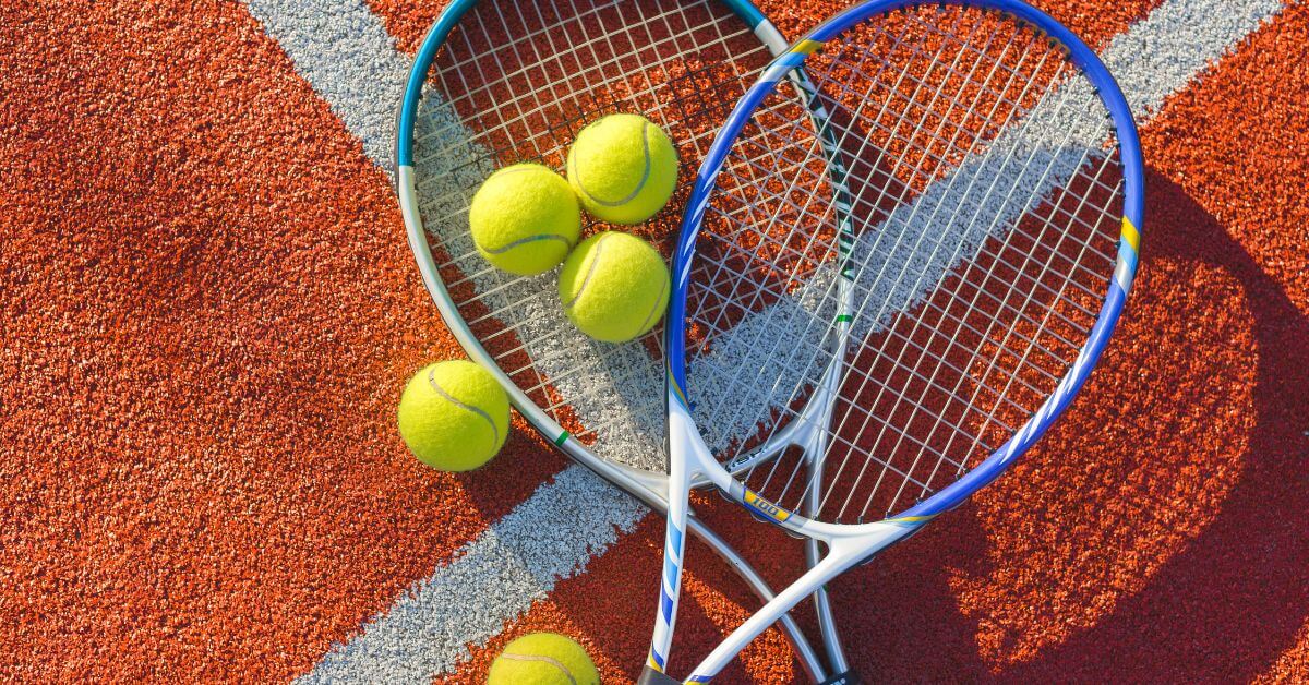 Two blue tennis rackets rest on an orange court alongside five tennis balls. The Best Tennis Rackets for Beginners should offer a balance of power and control for new players.