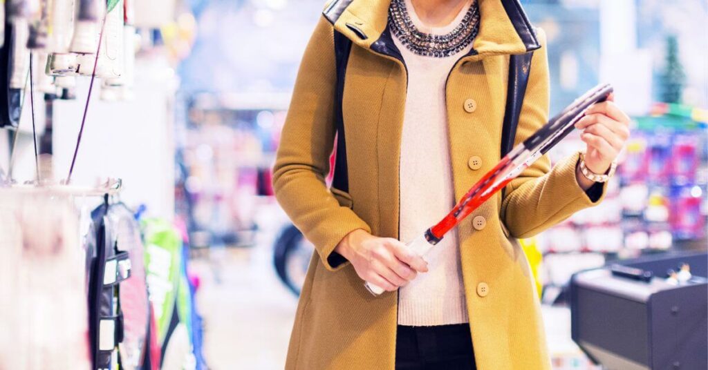 A woman in a mustard-colored coat browses a sporting goods store, holding a red and black tennis racket. She appears to be selecting from the Best Tennis Rackets for Beginners to start her tennis journey.