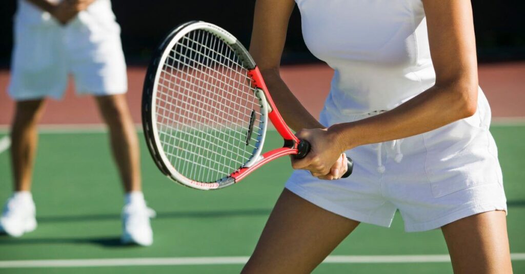 A female tennis player in white sportswear holds a red and black racket in a ready stance during a match. Finding the Best Tennis Rackets for Beginners is key to mastering powerful and precise shots.