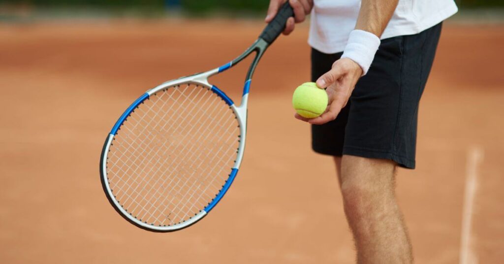 A tennis player prepares to serve on a clay court, holding a racket and a bright yellow ball. This scene highlights the importance of choosing the Best Tennis Rackets for Beginners to improve accuracy and control.