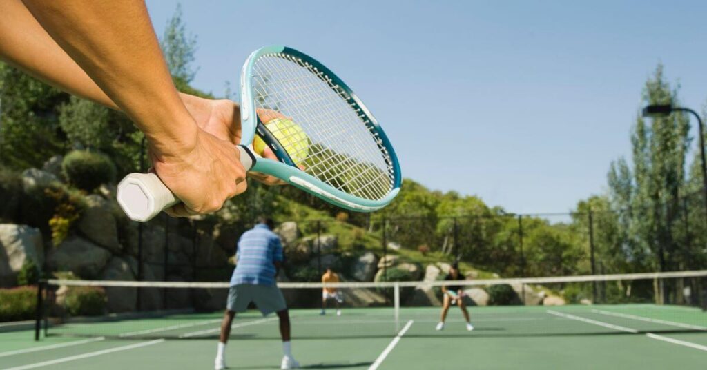 A close-up of a player gripping a light blue tennis racket, about to hit the ball during a doubles match on an outdoor court. The image emphasizes the need for the Best Tennis Rackets for Beginners to develop proper technique.