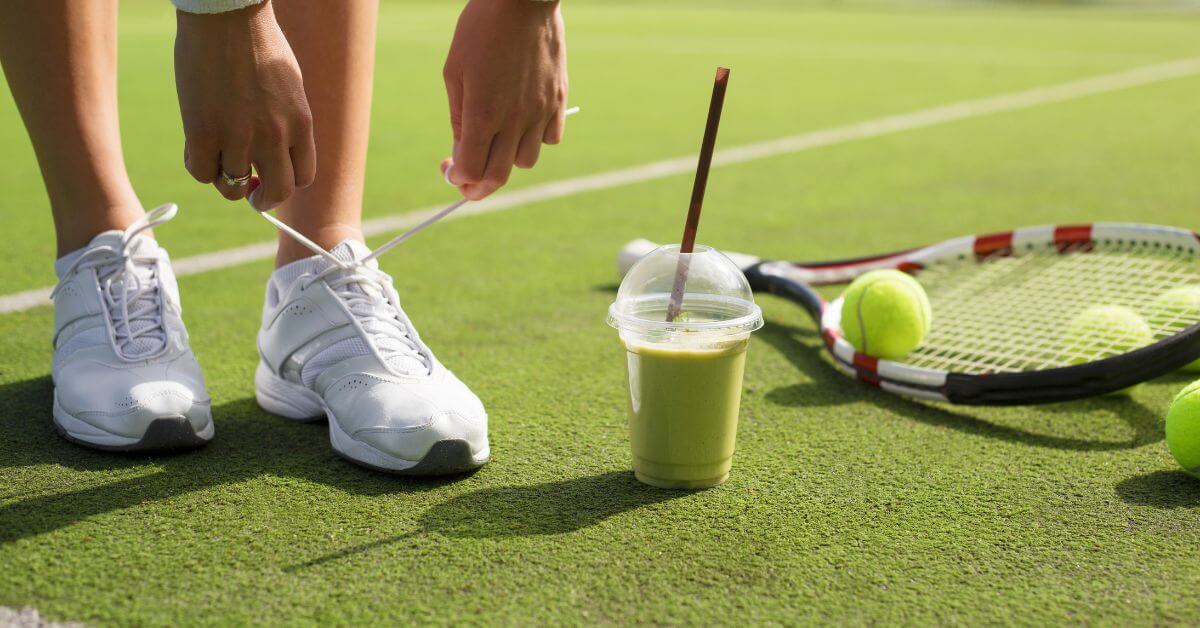 a person about to tie her women's tennis shoes on a green field with a green drink, green tennis ball, and a tennis racket on the side