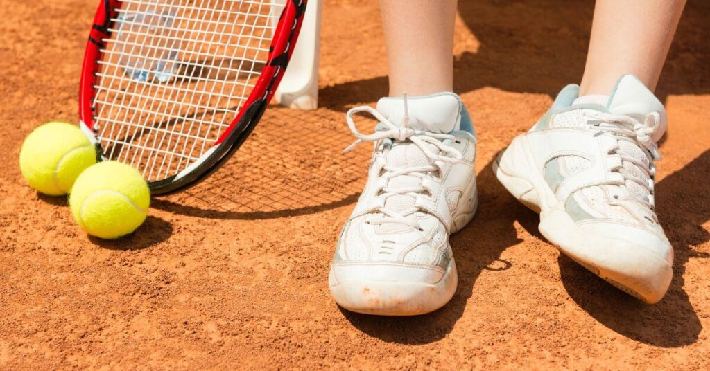 A close-up of a player's feet on a clay tennis court, wearing white sneakers with light blue accents. A tennis racket and two bright yellow tennis balls rest nearby, highlighting the best tennis shoes for women that offer stability and traction on clay surfaces.