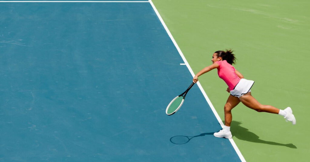 A female tennis player in a pink top and white skirt lunges forward on a blue and green hardcourt, swinging her racket. She wears white sneakers, demonstrating the best tennis shoes for women, built for agility and support during intense gameplay.