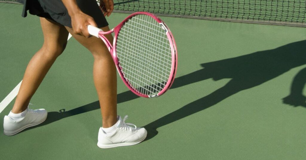 a person's legs on receiving stance with white tennis shoes while holding a red tennis racket on a green field