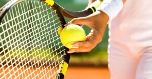 A female tennis player in a white outfit grips a yellow and black racket while holding a tennis ball against the strings, getting ready to serve. This demonstrates the proper technique for how to serve in tennis.