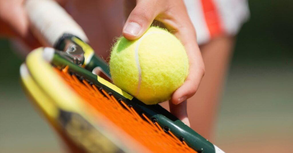 A detailed close-up of a player’s fingers positioning a fuzzy yellow tennis ball on an orange racket before serving. Understanding grip and ball placement is crucial when learning how to serve in tennis.