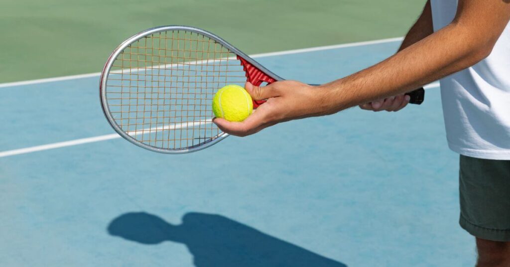 A close-up of a tennis player’s hands holding a racket and a tennis ball, preparing for a serve on a blue hardcourt. This image represents a key step in learning how to serve in tennis.