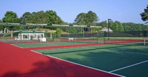 A well-maintained outdoor tennis facility with multiple hard courts featuring a green playing surface and red outer areas. The courts are enclosed by black fences, with a small pavilion and lush green trees in the background. This setting illustrates the structured and predictable playing conditions of hard courts in the ongoing "Clay vs Grass vs Hard Court" comparison.