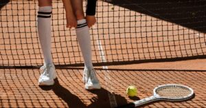 A hand extending a knee high sock with white shoes beside a tennis ball and racket, another way to know how to choose tennis shoes
