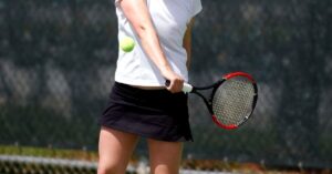 A person in a white shirt and black skirt prepares to hit a tennis ball mid-air with a racket on an outdoor court, perfect to start practicing tennis drills for beginners.